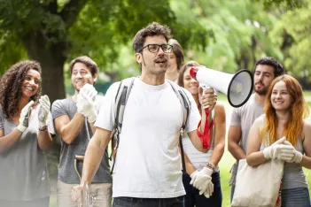 Young male volunteer giving speech on megaphone with other volunteers standing in background in park during garbage cleanup program