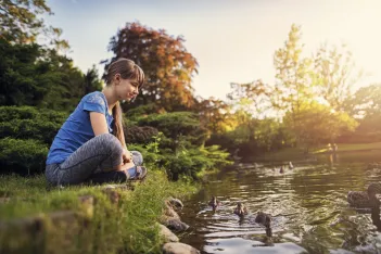 Girl sitting at the bank of a lake in city park looking at the ducks swimming in the pond.